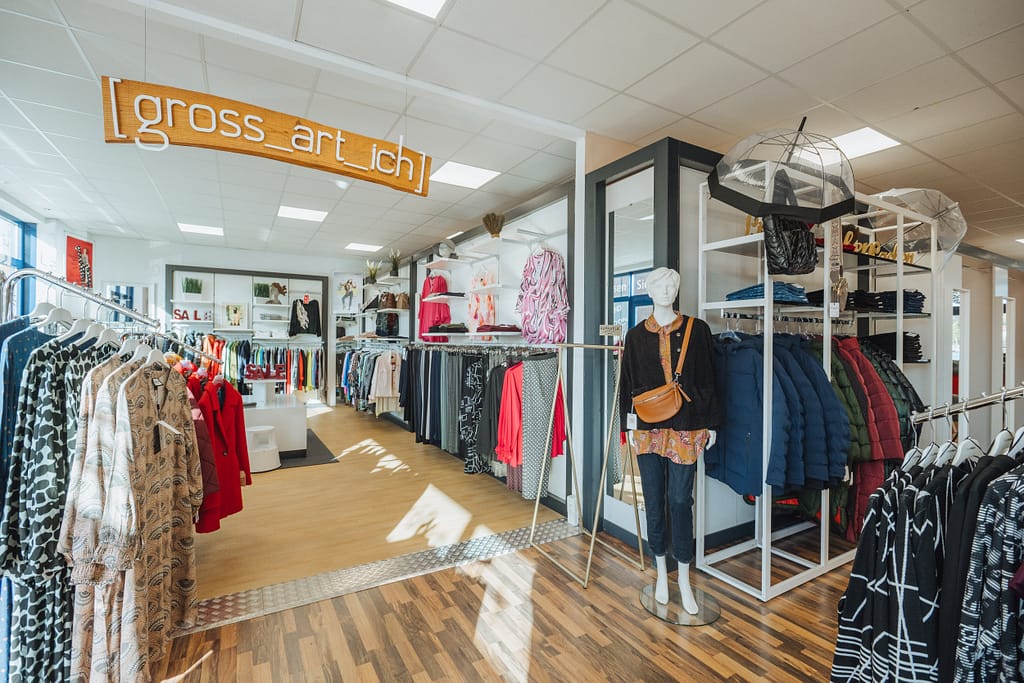 Im Modehaus "gross-art-ich" wird der linke Bereich des Fachgeschäfts für Grosse Grössen gezeigt. Ein großes Holzschild, weiß beschriftet mit dem Namen des Modehauses hängt von der Decke.